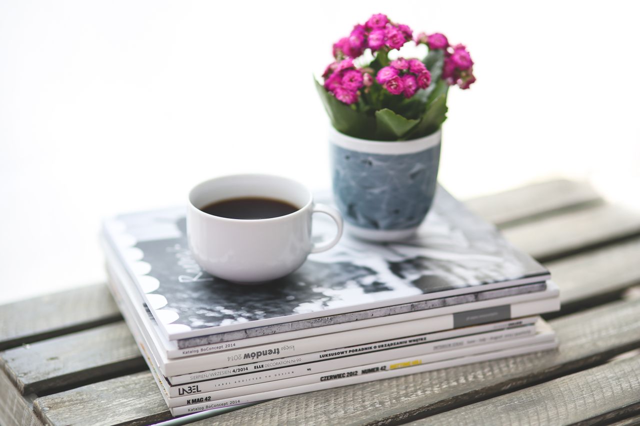 A cozy still life featuring coffee, magazines, and a pink flower pot on a rustic table.