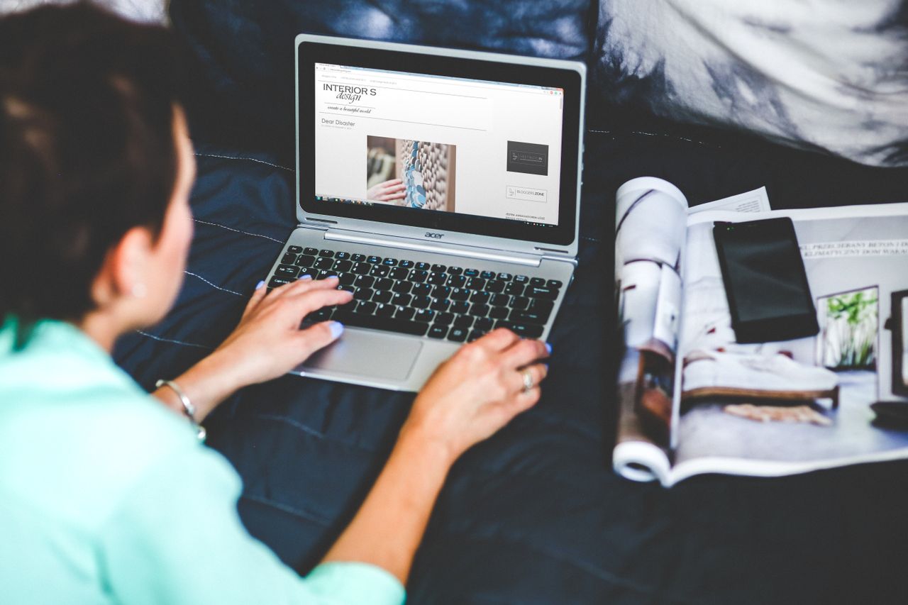 Welcome to Negative Marketing A woman on a bed browsing and blogging with a laptop and a magazine nearby.